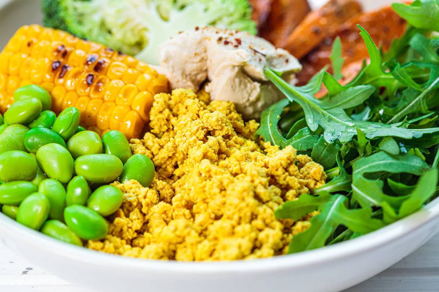 Plant-based bowl of tofu scramble, edamame, grilled corn, roasted carrots and rocket